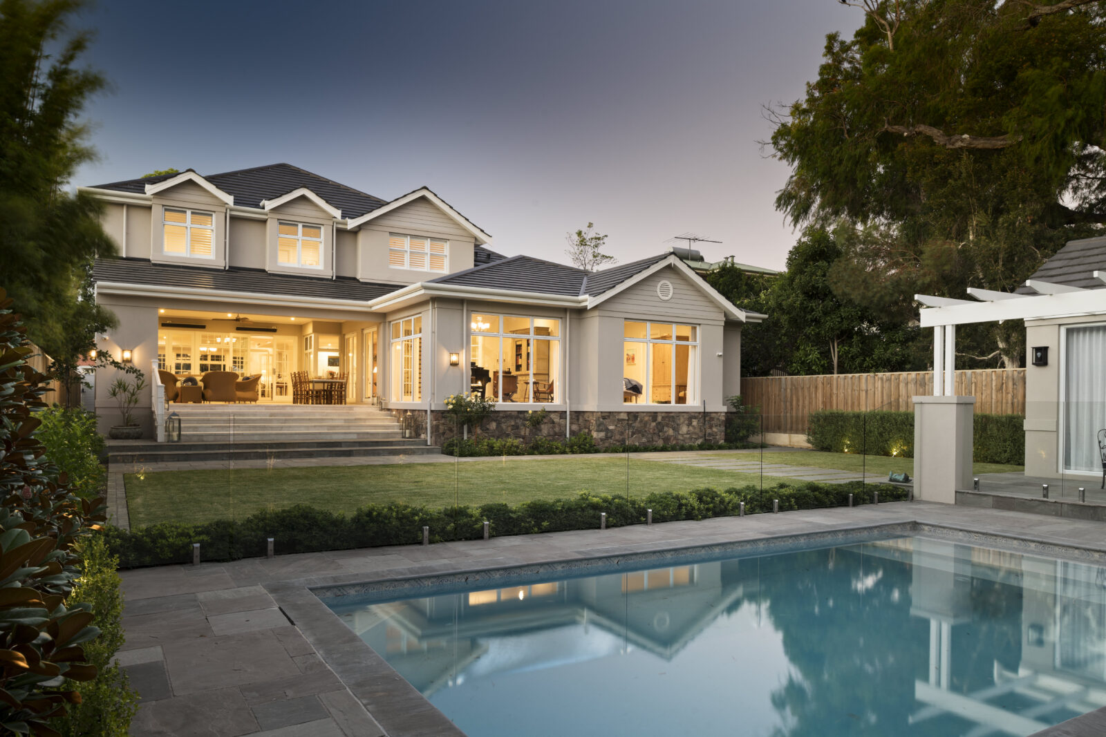 Outdoor luxury entertaining area with a sea blue pool in a high-end Hamptons home in Perth, featuring white trim awning, ambient warm lighting and glass pool framing surround. Part of a custom-built luxury home by Oswald Homes.