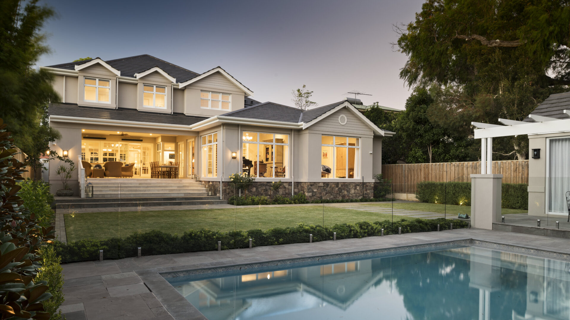 Outdoor luxury entertaining area with a sea blue pool in a high-end Hamptons home in Perth, featuring white trim awning, ambient warm lighting and glass pool framing surround. Part of a custom-built luxury home by Oswald Homes.
