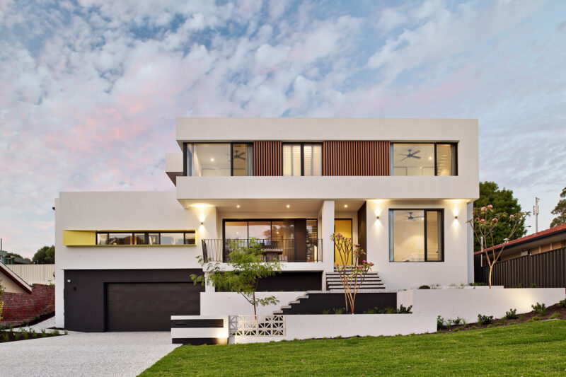 Two storey home facade at dusk with white render, timber accents and dark garage