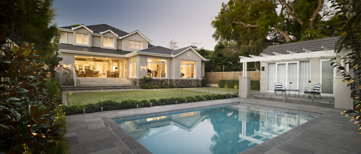Pool area and pool house located at the end of the garden of a double storey hamptons home with a glass pool fence and landscaped gardens