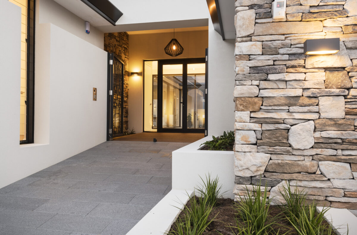 the walkway to the front entry of this custom home exudes warmth and luxury, Featuring a stone cladded feature wall and ambient lighting to welcome your guests. Built by luxury home builder, Oswald Homes.