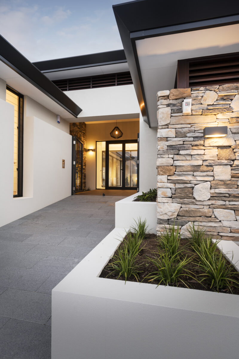 the walkway to the front entry of this custom home exudes warmth and luxury, Featuring a stone cladded feature wall and ambient lighting to welcome your guests. Built by luxury home builder, Oswald Homes.