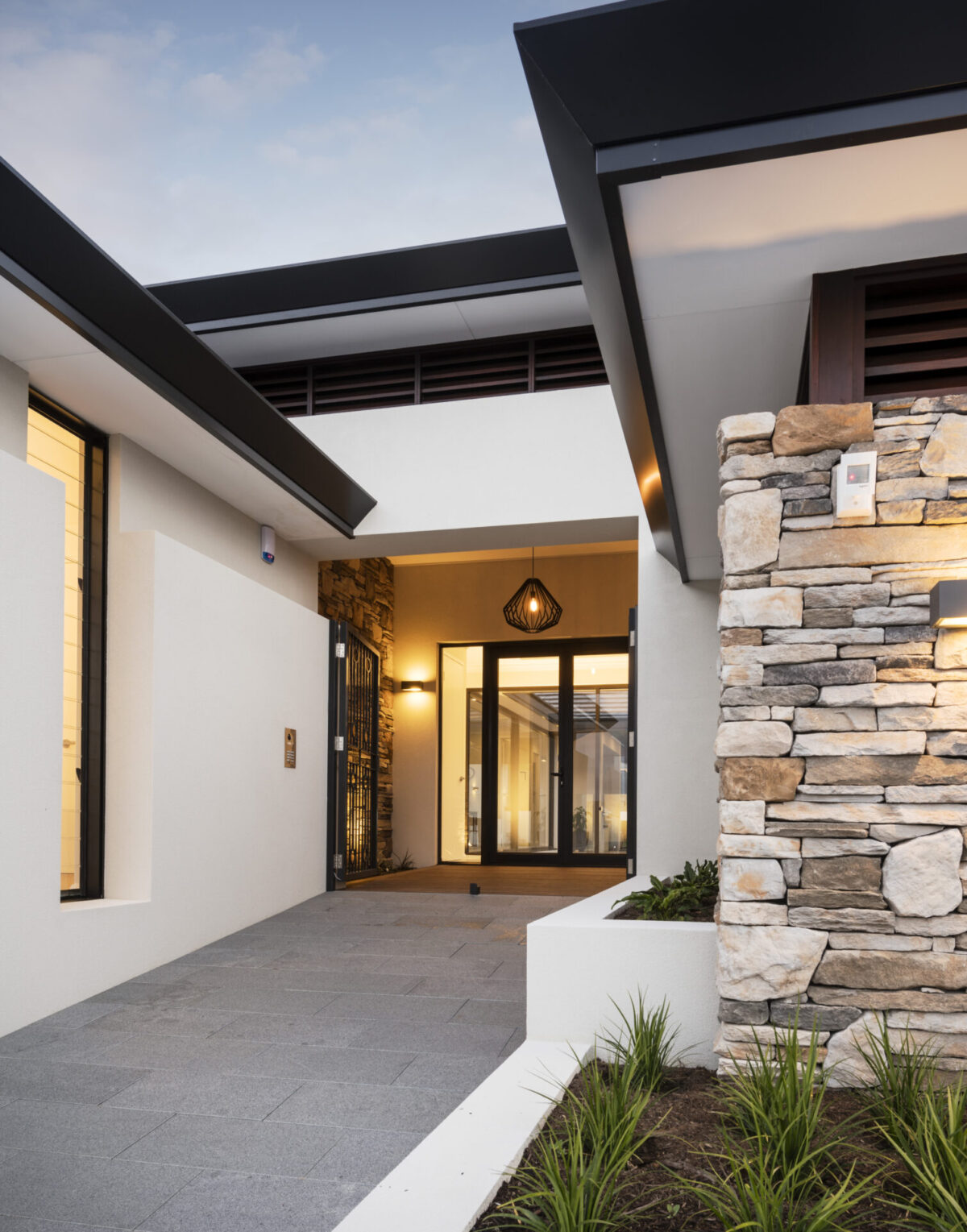 the walkway to the front entry of this custom home exudes warmth and luxury, Featuring a stone cladded feature wall and ambient lighting to welcome your guests. Built by luxury home builder, Oswald Homes.